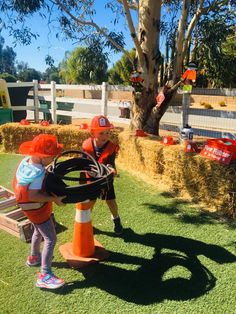 two children in orange safety vests are playing with a toy car on a fake grass field