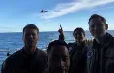 four men posing for a photo on a ship in the ocean with an airplane flying overhead