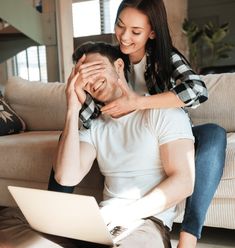 a man and woman sitting on a couch looking at a laptop computer while they laugh