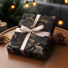 a present wrapped in black and gold paper with a white ribbon on a table next to pine cones