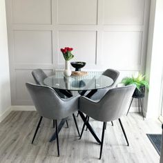 a glass table with four grey chairs in front of white paneled walls and wood flooring