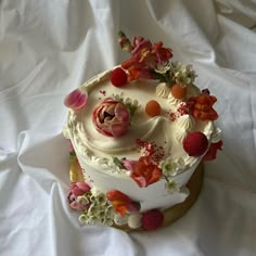 a white cake with fruit and flowers on it sitting on a tablecloth covered surface