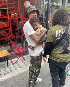 a man holding a baby in his arms while standing next to a woman on the sidewalk