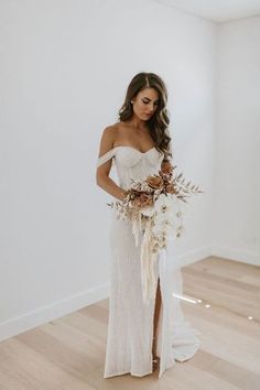 a woman standing in a room holding a bouquet and wearing a white dress with an off the shoulder neckline