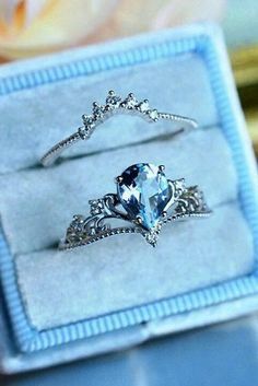 two engagement rings in a box on a blue surface with flowers behind them and the ring has an aquamarine colored stone surrounded by silver fili