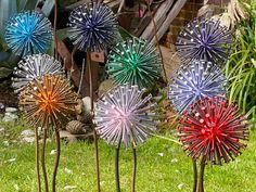 four colorful metal flowers in the grass near a brick wall and building with plants behind them