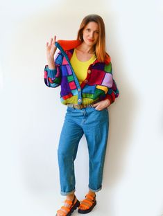 a woman standing in front of a white wall wearing blue jeans and a colorful jacket