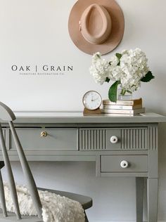 a desk with a hat on top and flowers in a vase sitting on it next to a chair