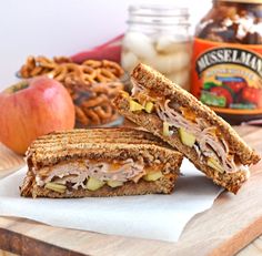 a sandwich cut in half sitting on top of a cutting board next to apples and pretzels