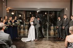 a bride and groom kissing in front of their wedding party at the winery with wine casks behind them