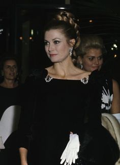 a woman in a black dress with white gloves on her hand and other people behind her