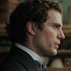 a man with curly hair wearing a suit and tie looking off to the side in front of bookshelves