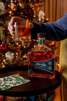 a person holding a glass near a bottle of alcohol on a table in front of a christmas tree