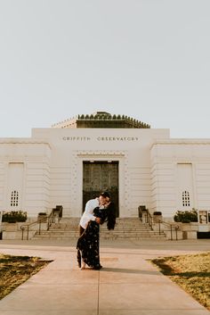 an engaged couple kissing in front of a large white building with steps leading up to it