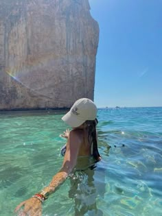 a woman swimming in the ocean next to a large rock formation with a sun hat on