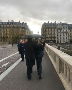 two people walking on a bridge with an umbrella over their heads and buildings in the background