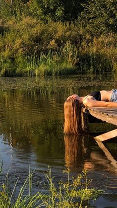 a woman laying on top of a wooden dock next to a body of water with trees in the background