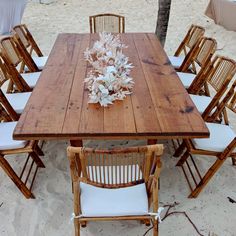 a wooden table with white chairs around it on the sand near some trees and water