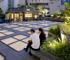 two people sitting on a bench in the middle of a courtyard with plants and potted trees