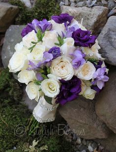 a bridal bouquet with purple and white flowers is sitting on the ground next to rocks