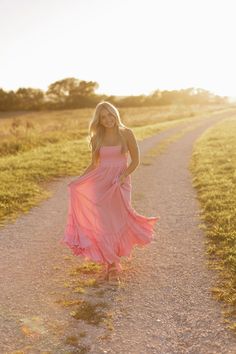 a woman in a pink dress is walking down a dirt road with her hands on her hips