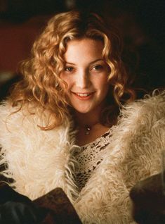 a woman with curly hair wearing a white fur coat and smiling at the camera while sitting on a couch