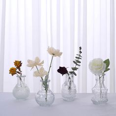 three clear vases with white and yellow flowers in them on a table next to sheer curtains