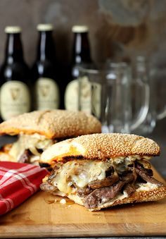 two sandwiches with meat and cheese on a cutting board next to bottles of beer in the background