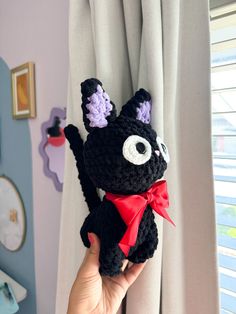 a hand holding up a black crocheted cat with purple ears and tail, wearing a red bow