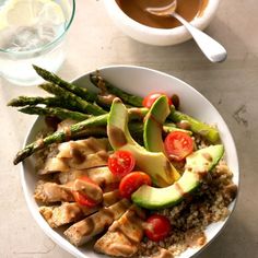 a white bowl filled with chicken, asparagus and tomatoes next to a cup of coffee