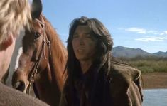 a man with long hair standing next to a horse and looking at another person's face