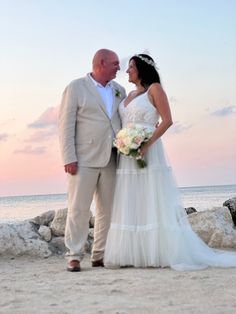 a bride and groom standing on the beach at sunset