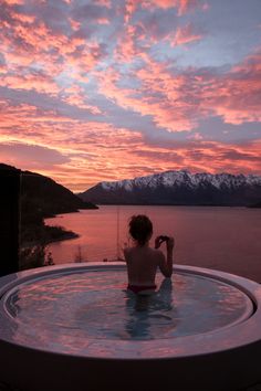 a woman sitting in a hot tub with the sun setting behind her and mountains in the distance