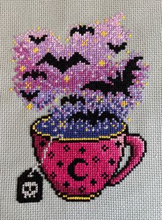 a cross - stitch picture of a coffee cup with purple flowers in it