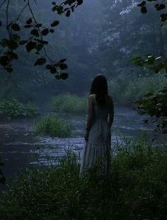 a woman standing in front of a body of water with trees and bushes around her