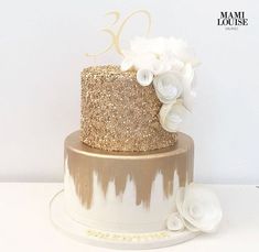 a gold and white cake with flowers on top is displayed in front of a white wall