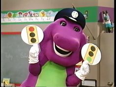 an inflatable purple dinosaur is holding up two yellow traffic lights while wearing a baseball cap and green shirt