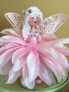 a pink and white flower fairy sitting on top of a green table next to a yellow wall