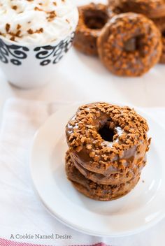 three chocolate donuts on a white plate next to a cup of whipped cream and other desserts