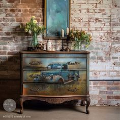 an old dresser with flowers and candles on top in front of a brick wall, next to a painting