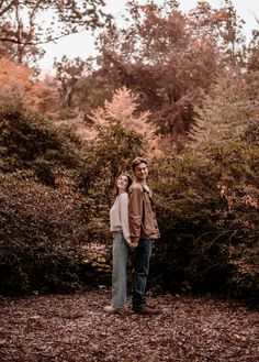 a man and woman standing next to each other in front of some trees with leaves on the ground