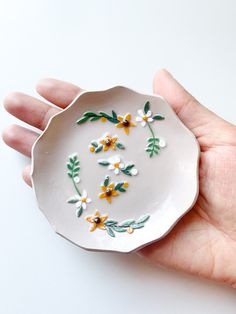 a hand holding a small white plate with yellow and white flowers on it's rim