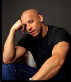 a bald man sitting on top of a bed next to a white pillow and wearing a black t - shirt