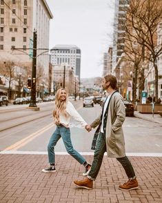 a man and woman holding hands while walking down the street with buildings in the background