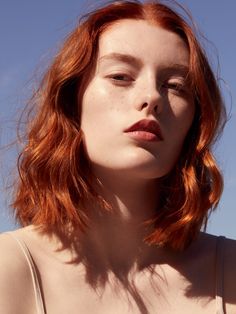 a woman with red hair wearing a tank top and looking off into the distance, against a blue sky