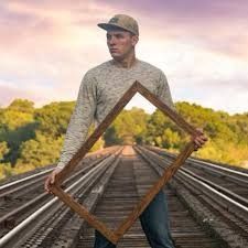 a man standing on train tracks holding up a framed photo with trees in the background