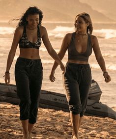 two young women walking on the beach with a boat in the background and one holding her hand