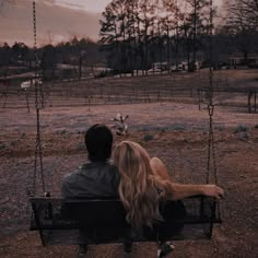 a man and woman sitting on a swing in the middle of a field at sunset