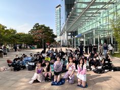 a group of people sitting on the ground in front of a glass building with lots of windows