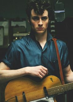 a young man holding an acoustic guitar in his right hand and looking at the camera